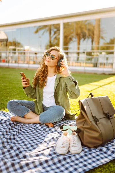 Kablosuz kulaklığı ve telefonu olan genç bir kadın güneşli bir parkta, palmiye ağaçlarıyla çevrili bir battaniyenin üzerinde oturuyor. Güzel kadın eğleniyor, müzik dinliyor ve dışarıda dinleniyor. Yaşam biçimi.