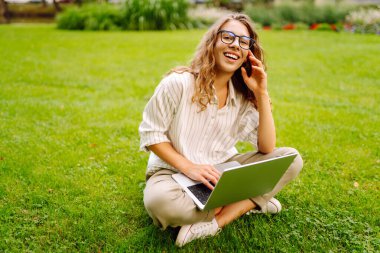 Neşeli bir kadın güneşli bir parkta çimlerde dizüstü bilgisayarla oturuyor. Gün batımında dizüstü bilgisayarda çalışan gözlüklü bir kadın serbest yazar. Uzaktan çalışma, öğrenme ve rahatlama kavramı.