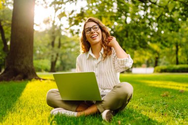 Neşeli bir kadın güneşli bir parkta çimlerde dizüstü bilgisayarla oturuyor. Gün batımında dizüstü bilgisayarda çalışan gözlüklü bir kadın serbest yazar. Uzaktan çalışma, öğrenme ve rahatlama kavramı.