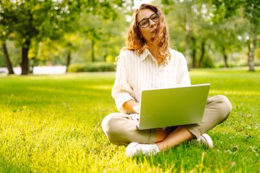 Neşeli bir kadın güneşli bir parkta çimlerde dizüstü bilgisayarla oturuyor. Gün batımında dizüstü bilgisayarda çalışan gözlüklü bir kadın serbest yazar. Uzaktan çalışma, öğrenme ve rahatlama kavramı.