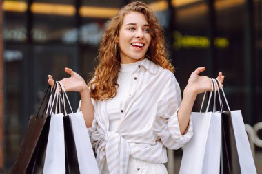 Alışveriş torbalı mutlu bir kadın güneşli bir günde büyük bir alışveriş merkezinin yanında durur. Genç bir kadın alışverişten sonra dışarıda vakit geçirmekten hoşlanır. Alışveriş konsepti, tüketim ve indirimler.