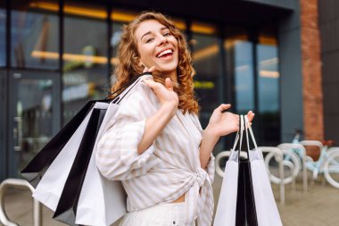 Alışveriş torbalı mutlu bir kadın güneşli bir günde büyük bir alışveriş merkezinin yanında durur. Genç bir kadın alışverişten sonra dışarıda vakit geçirmekten hoşlanır. Alışveriş konsepti, tüketim ve indirimler.