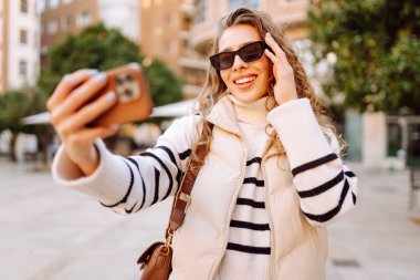 Güneş gözlüklü neşeli kadın, güneşli şehir sokaklarında dolaşırken telefonla konuşuyor. Güzel turistler şehir havasını seviyorlar ve dışarıda telefonda selfie çekiyorlar. Turizm, blog ve yürüme kavramları.