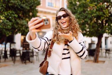 Güneş gözlüklü neşeli kadın, güneşli şehir sokaklarında dolaşırken telefonla konuşuyor. Güzel turistler şehir havasını seviyorlar ve dışarıda telefonda selfie çekiyorlar. Turizm, blog ve yürüme kavramları.