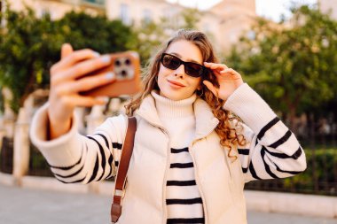 Güneş gözlüklü neşeli kadın, güneşli şehir sokaklarında dolaşırken telefonla konuşuyor. Güzel turistler şehir havasını seviyorlar ve dışarıda telefonda selfie çekiyorlar. Turizm, blog ve yürüme kavramları.