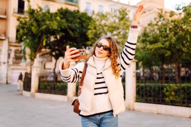 Güneş gözlüklü neşeli kadın, güneşli şehir sokaklarında dolaşırken telefonla konuşuyor. Güzel turistler şehir havasını seviyorlar ve dışarıda telefonda selfie çekiyorlar. Turizm, blog ve yürüme kavramları.
