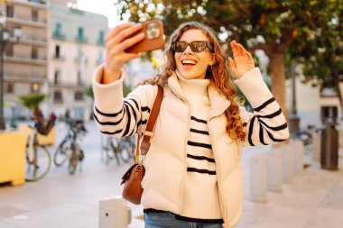 Güneş gözlüklü neşeli kadın, güneşli şehir sokaklarında dolaşırken telefonla konuşuyor. Güzel turistler şehir havasını seviyorlar ve dışarıda telefonda selfie çekiyorlar. Turizm, blog ve yürüme kavramları.