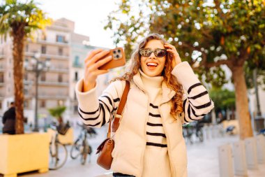 Güneş gözlüklü neşeli kadın, güneşli şehir sokaklarında dolaşırken telefonla konuşuyor. Güzel turistler şehir havasını seviyorlar ve dışarıda telefonda selfie çekiyorlar. Turizm, blog ve yürüme kavramları.