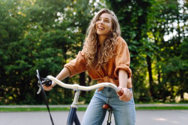 Neşeli bir kadın yeşil bir parkta bir yol boyunca bisiklet sürüyor. Güzel bir kadın güneşli bir günde bisiklet sürmekten hoşlanır. Kavram: rahatlama, yürüyüş, aktif yaşam tarzı.