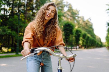 Neşeli bir kadın yeşil bir parkta bir yol boyunca bisiklet sürüyor. Güzel bir kadın güneşli bir günde bisiklet sürmekten hoşlanır. Kavram: rahatlama, yürüyüş, aktif yaşam tarzı.
