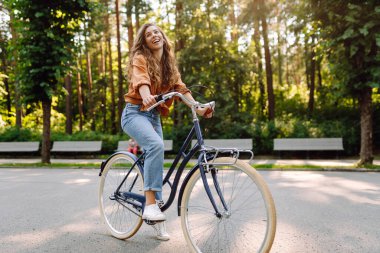 Neşeli bir kadın yeşil bir parkta bir yol boyunca bisiklet sürüyor. Güzel bir kadın güneşli bir günde bisiklet sürmekten hoşlanır. Kavram: rahatlama, yürüyüş, aktif yaşam tarzı.