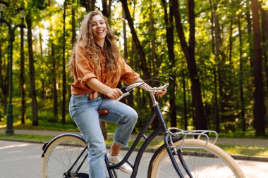 Neşeli bir kadın yeşil bir parkta bir yol boyunca bisiklet sürüyor. Güzel bir kadın güneşli bir günde bisiklet sürmekten hoşlanır. Kavram: rahatlama, yürüyüş, aktif yaşam tarzı.