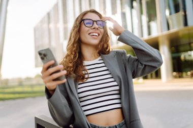 Gözlük takan ve akıllı telefon kullanan neşeli bir kadın modern bir binanın yanında duruyor. Güzel iş kadını telefonuna mesaj atıyor ve güneşli günün tadını çıkarıyor. Modernlik kavramı, kentsel.