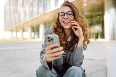 Gözlük takan ve akıllı telefon kullanan neşeli bir kadın modern bir binanın yanında duruyor. Güzel iş kadını telefonuna mesaj atıyor ve güneşli günün tadını çıkarıyor. Modernlik kavramı, kentsel.