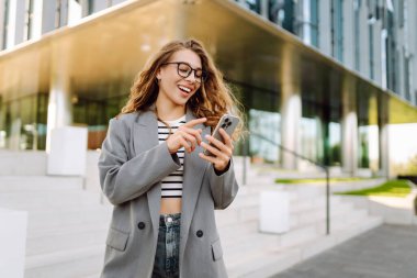 Gözlük takan ve akıllı telefon kullanan neşeli bir kadın modern bir binanın yanında duruyor. Güzel iş kadını telefonuna mesaj atıyor ve güneşli günün tadını çıkarıyor. Modernlik kavramı, kentsel.