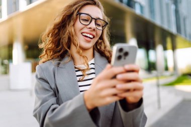 Gözlük takan ve akıllı telefon kullanan neşeli bir kadın modern bir binanın yanında duruyor. Güzel iş kadını telefonuna mesaj atıyor ve güneşli günün tadını çıkarıyor. Modernlik kavramı, kentsel.