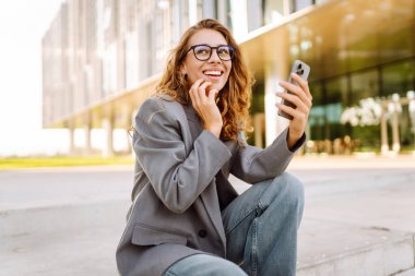 Gözlük takan ve akıllı telefon kullanan neşeli bir kadın modern bir binanın yanında duruyor. Güzel iş kadını telefonuna mesaj atıyor ve güneşli günün tadını çıkarıyor. Modernlik kavramı, kentsel.