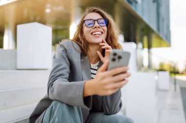 Gözlük takan ve akıllı telefon kullanan neşeli bir kadın modern bir binanın yanında duruyor. Güzel iş kadını telefonuna mesaj atıyor ve güneşli günün tadını çıkarıyor. Modernlik kavramı, kentsel.