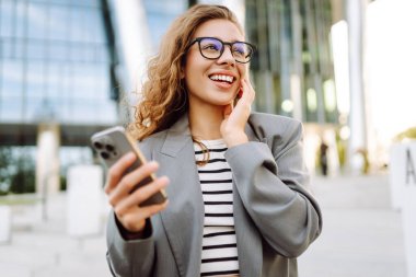 Gözlük takan ve akıllı telefon kullanan neşeli bir kadın modern bir binanın yanında duruyor. Güzel iş kadını telefonuna mesaj atıyor ve güneşli günün tadını çıkarıyor. Modernlik kavramı, kentsel.