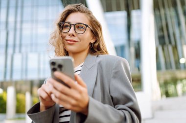 Gözlük takan ve akıllı telefon kullanan neşeli bir kadın modern bir binanın yanında duruyor. Güzel iş kadını telefonuna mesaj atıyor ve güneşli günün tadını çıkarıyor. Modernlik kavramı, kentsel.