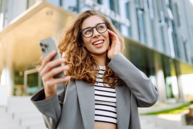 Gözlük takan ve akıllı telefon kullanan neşeli bir kadın modern bir binanın yanında duruyor. Güzel iş kadını telefonuna mesaj atıyor ve güneşli günün tadını çıkarıyor. Modernlik kavramı, kentsel.