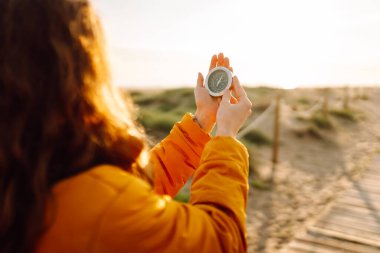 Parlak ceketli ve sırt çantalı genç bir yürüyüşçü bir pusula tutuyor ve deniz manzarasına bakıyor. Yürüyüşçü gün batımında yürüyüş yolunda dururken bir yön seçer. Macera kavramı, navigasyon.