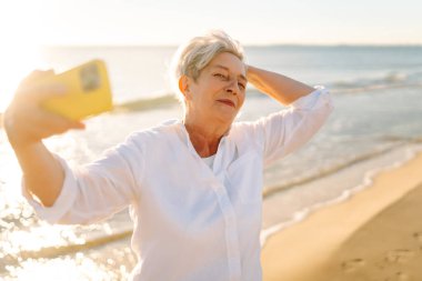 Neşeli yaşlı kadın sahilde duruyor, deniz manzarasına tepeden bakan bir telefonla selfie çekiyor. Mutlu emekliler dışarıda eğleniyor ve blog yazıyor. Blog yazma kavramı, özgürlük. Mutlu emeklilik.