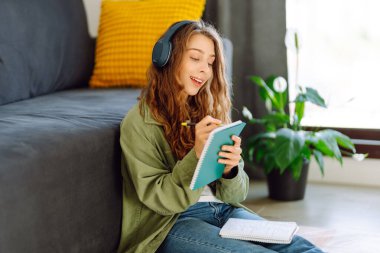 Kablosuz kulaklık takan genç bir kadın rahat oturma odasında oturur ve not defterine not alır. Güzel bir kadın kitap okumayı ya da not almayı sever. Eğitim, rahatlama ve konfor kavramı.