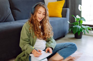 Kablosuz kulaklık takan genç bir kadın rahat oturma odasında oturur ve not defterine not alır. Güzel bir kadın kitap okumayı ya da not almayı sever. Eğitim, rahatlama ve konfor kavramı.