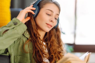 Kablosuz kulaklık takan genç bir kadın rahat oturma odasında oturur ve not defterine not alır. Güzel bir kadın kitap okumayı ya da not almayı sever. Eğitim, rahatlama ve konfor kavramı.