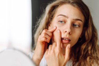 Küçük aynada cildini dikkatlice inceleyen genç bir kadının yakın çekimi. Sivilceli güzel bir kadın rahat bir odada yüzüne dokunuyor. Doğallık ve güzellik kavramı, dermatoloji. Sivilce.