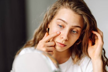 Küçük aynada cildini dikkatlice inceleyen genç bir kadının yakın çekimi. Sivilceli güzel bir kadın rahat bir odada yüzüne dokunuyor. Doğallık ve güzellik kavramı, dermatoloji. Sivilce.