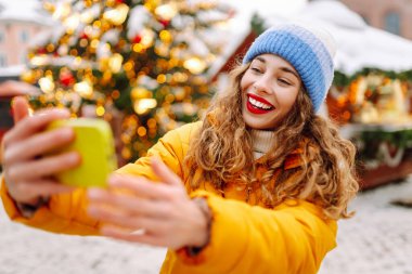 Kırmızı rujlu neşeli kadın kış günü canlı fuarda selfie çekiyor. Yeni yıl tatili boyunca parlak ceket blogları ve Noel ağacı yakınında gezinen bir kadın. Bloglama ve kutlama kavramları.