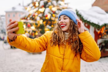 Kırmızı rujlu neşeli kadın kış günü canlı fuarda selfie çekiyor. Yeni yıl tatili boyunca parlak ceket blogları ve Noel ağacı yakınında gezinen bir kadın. Bloglama ve kutlama kavramları.