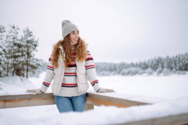 Rahat bir kazak ve şapka giymiş genç bir kadın etrafı ormanla çevrili karla kaplı bir göle bakıyor. Bir yürüyüşçü yürüyüş parkurunda karlı bir günün tadını çıkarır. Mevsimlik, rahatlama ve macera kavramları.
