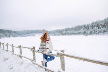 Rahat bir kazak ve şapka giymiş genç bir kadın etrafı ormanla çevrili karla kaplı bir göle bakıyor. Bir yürüyüşçü yürüyüş parkurunda karlı bir günün tadını çıkarır. Mevsimlik, rahatlama ve macera kavramları.