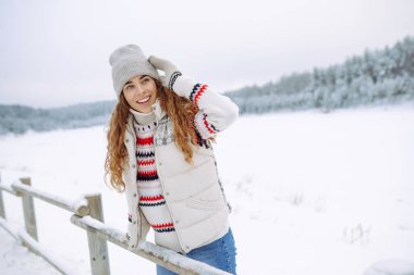 Rahat bir kazak ve şapka giymiş genç bir kadın etrafı ormanla çevrili karla kaplı bir göle bakıyor. Bir yürüyüşçü yürüyüş parkurunda karlı bir günün tadını çıkarır. Mevsimlik, rahatlama ve macera kavramları.
