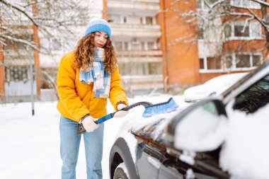 Parlak ceketli ve şapkalı mutlu kadın kış sabahı arabasındaki karı temizliyor. Karlı bir günde özel bir fırça temizleme arabası olan kadın bir sürücünün yakın çekimi. Kavram: Mevsimlik, kentsellik, yaşam tarzı.