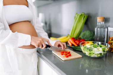 Hamile kadın modern mutfakta duruyor, taze sebze ve meyve doğruyor. Bebek bekleyen genç bir kadın sağlıklı yemek hazırlıyor ve sıcak odada güneşli bir günün tadını çıkarıyor. Konsept sağlık, bakım, keyif.