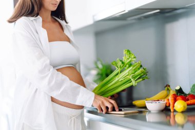Hamile kadın modern mutfakta duruyor, taze sebze ve meyve doğruyor. Bebek bekleyen genç bir kadın sağlıklı yemek hazırlıyor ve sıcak odada güneşli bir günün tadını çıkarıyor. Konsept sağlık, bakım, keyif.