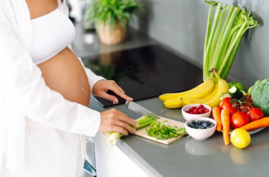 Hamile kadın modern mutfakta duruyor, taze sebze ve meyve doğruyor. Bebek bekleyen genç bir kadın sağlıklı yemek hazırlıyor ve sıcak odada güneşli bir günün tadını çıkarıyor. Konsept sağlık, bakım, keyif.