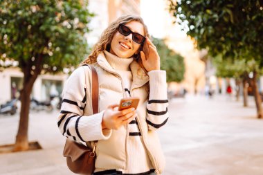 Güneş gözlüklü genç bir kadının portresi. Şehir caddesinde dururken telefonla mesajlaşıyor. Neşeli bir kadın güneşli bir günde şehrin mimarisinden zevk alır. Turizm, blog yazma ve yürüme kavramı.