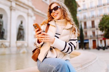 Güneş gözlüklü genç bir kadının portresi. Şehir caddesinde dururken telefonla mesajlaşıyor. Neşeli bir kadın güneşli bir günde şehrin mimarisinden zevk alır. Turizm, blog yazma ve yürüme kavramı.