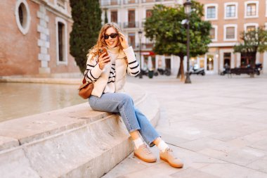 Güneş gözlüklü genç bir kadının portresi. Şehir caddesinde dururken telefonla mesajlaşıyor. Neşeli bir kadın güneşli bir günde şehrin mimarisinden zevk alır. Turizm, blog yazma ve yürüme kavramı.