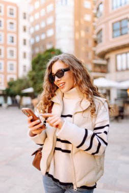 Güneş gözlüklü genç bir kadının portresi. Şehir caddesinde dururken telefonla mesajlaşıyor. Neşeli bir kadın güneşli bir günde şehrin mimarisinden zevk alır. Turizm, blog yazma ve yürüme kavramı.