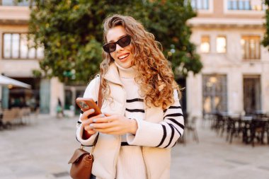 Güneş gözlüklü genç bir kadının portresi. Şehir caddesinde dururken telefonla mesajlaşıyor. Neşeli bir kadın güneşli bir günde şehrin mimarisinden zevk alır. Turizm, blog yazma ve yürüme kavramı.