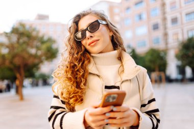 Güneş gözlüklü genç bir kadının portresi. Şehir caddesinde dururken telefonla mesajlaşıyor. Neşeli bir kadın güneşli bir günde şehrin mimarisinden zevk alır. Turizm, blog yazma ve yürüme kavramı.