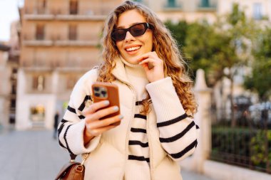 Güneş gözlüklü genç bir kadının portresi. Şehir caddesinde dururken telefonla mesajlaşıyor. Neşeli bir kadın güneşli bir günde şehrin mimarisinden zevk alır. Turizm, blog yazma ve yürüme kavramı.