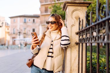 Güneş gözlüklü genç bir kadının portresi. Şehir caddesinde dururken telefonla mesajlaşıyor. Neşeli bir kadın güneşli bir günde şehrin mimarisinden zevk alır. Turizm, blog yazma ve yürüme kavramı.