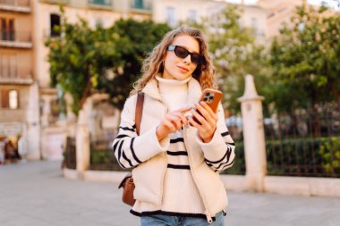 Güneş gözlüklü genç bir kadının portresi. Şehir caddesinde dururken telefonla mesajlaşıyor. Neşeli bir kadın güneşli bir günde şehrin mimarisinden zevk alır. Turizm, blog yazma ve yürüme kavramı.